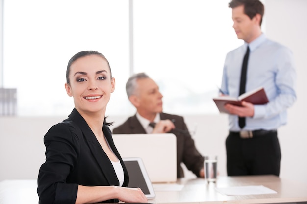 Happy to be a part of team. Attractive young woman in formalwear looking over shoulder and smiling while sitting at the table with two men talking on background