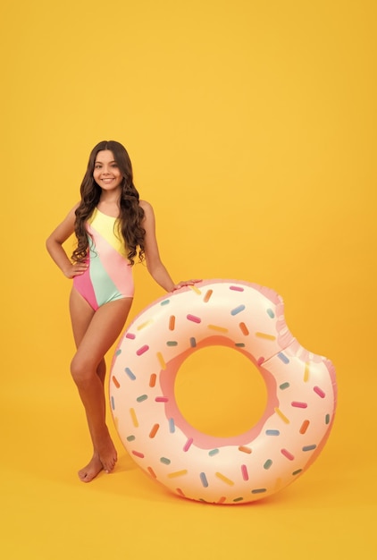 Happy beach teen girl with curly hair in swimsuit with doughnut inflatable ring for pool party fun on summer vacation on yellow background beach accessory