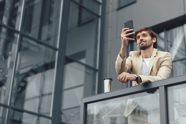 Felice barbuto uomo d'affari in tuta e smartphone wireless avente videoconferenza su smartphone all'aperto