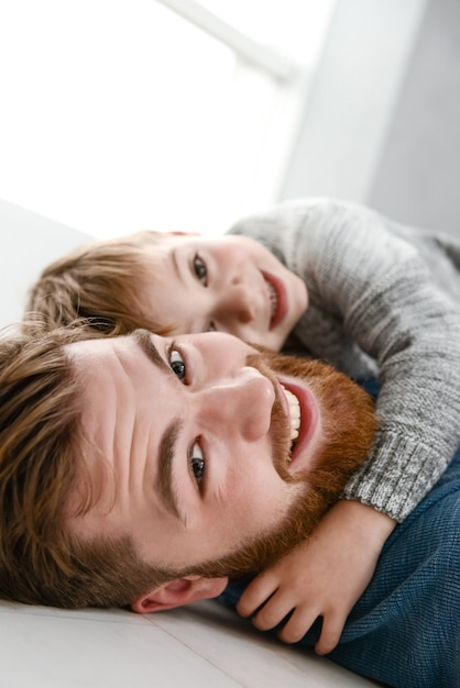 屋内の部屋で彼の小さなかわいい息子と遊んで青いセーターを着た幸せなひげを生やした父