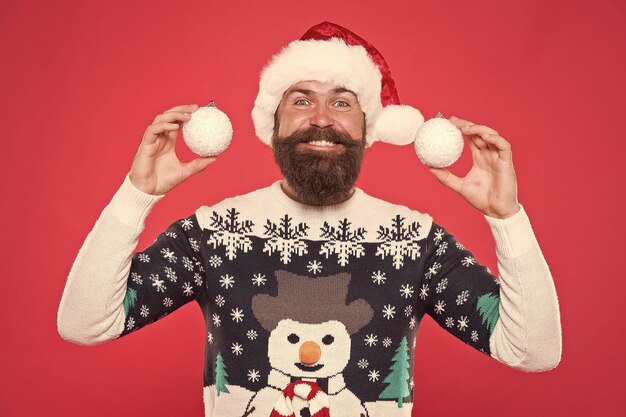 Foto felice ragazzo barbuto in caldo maglione lavorato a maglia e cappello di babbo natale tiene palle di neve decorative sull'albero celebra le vacanze invernali di natale capodanno arredamento