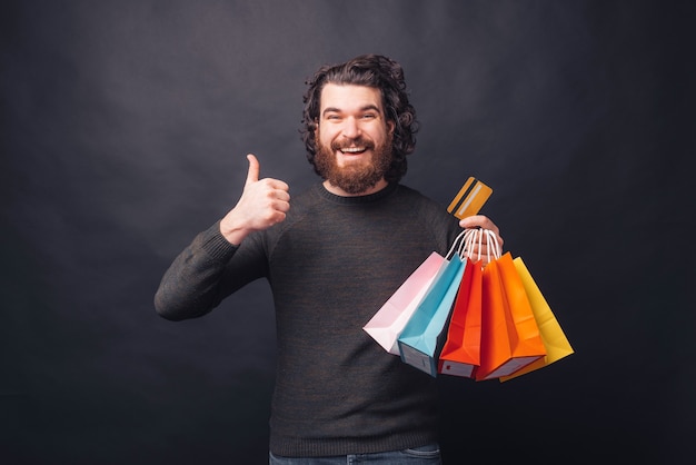 Happy bearded hipster man showing thumb up and holding shopping bags and credit card