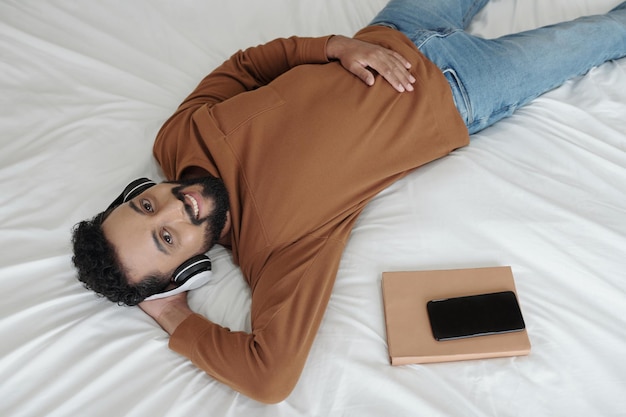 Foto uomo barbuto felice in abiti casual che si rilassa sul letto ascoltando musica in cuffia e alzando lo sguardo...