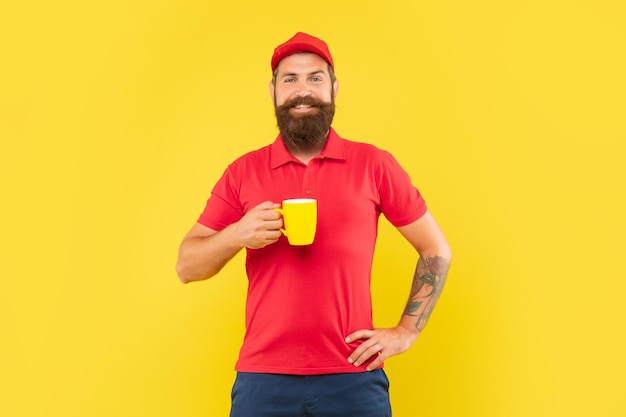 Happy bearded man in casual red cap and tshirt holding mug yellow background coffee