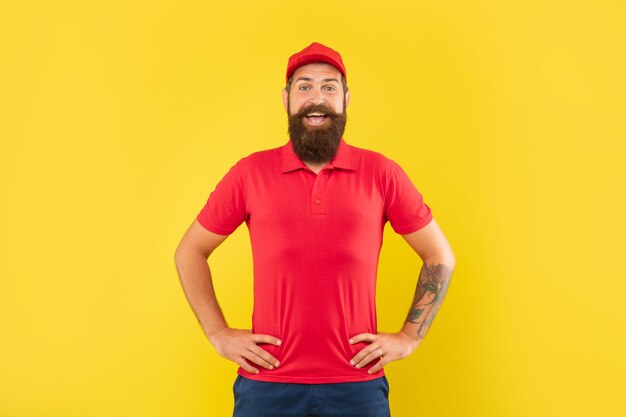 캐주얼 빨간 모자와 문신된 팔 akimbo 노란색 배경 남자와 서 있는 tshirt에서 행복 한 수염된 남자