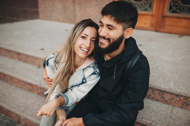 Happy bearded man and cheerful woman embracing while sitting on steps on street