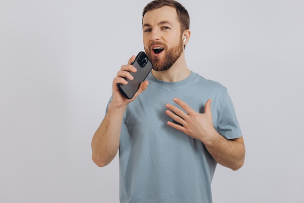 Felice uomo barbuto che balla con gli auricolari ascoltando musica in cuffia tenendo il telefono cellulare e sorridendo compiaciuto in piedi su sfondo bianco