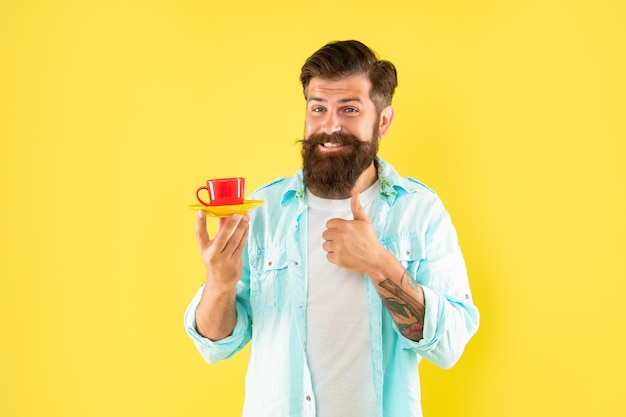 幸せなひげを生やした男は黄色の背景のコーヒー カップに親指を現してモーニング コーヒーを飲む