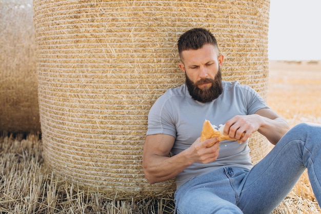 Happy bearded man farmer in jeans holding fresh fragrant bread and sitting near the bale around sunny sun field