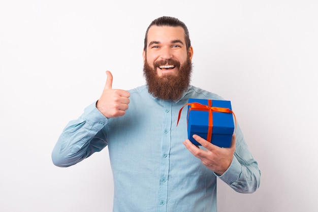 Happy bearded man is showing thumb up while holding a gift box.