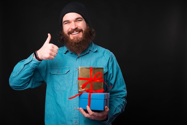 Счастливый бородатый мужчина улыбается в камеру и держит большой палец вверх, и некоторые подарки показывают, что ему нравятся подарки