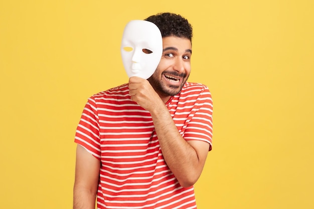 Happy bearded man removing white mask from face showing his smiling expression, good mood, pretending to be another person. Indoor studio shot isolated on yellow background