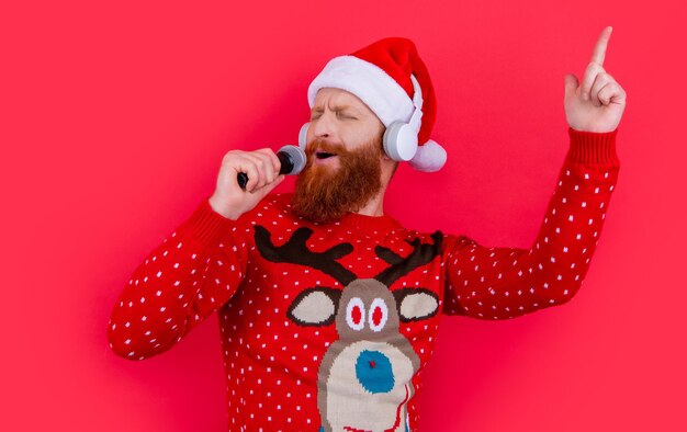 Happy bearded man in santa hat singing christmas song pointing\
finger up studio isolated on red