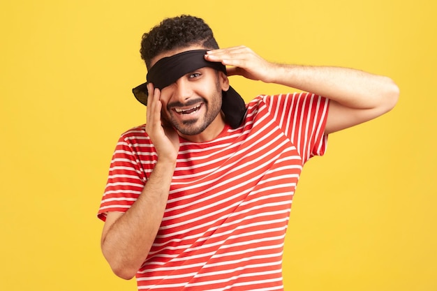 Happy bearded man in striped red tshirt taking off blindfold\
from eyes to look at his surprise looking at camera with toothy\
smile indoor studio shot isolated on yellow background