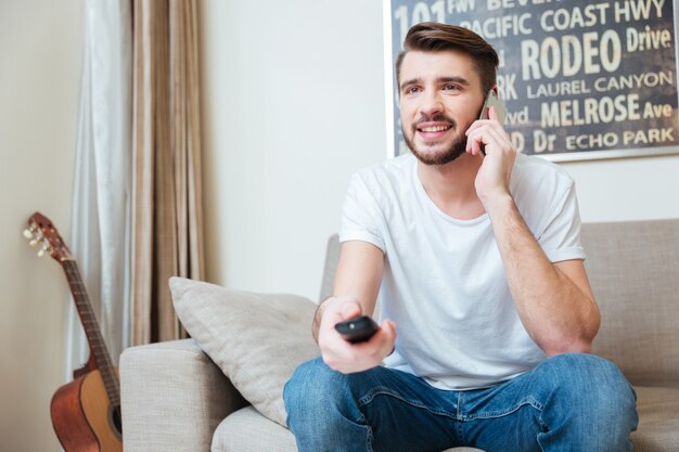 Happy bearded man using remote control and talking on cell phone on sofa at home