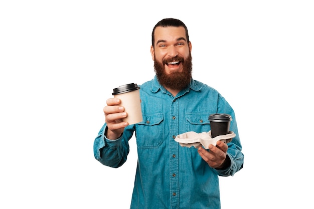 파란색 셔츠를 입은 행복한 수염 난 남자가 테이크아웃 커피 컵 두 개를 들고 있다