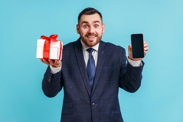 Happy bearded man wearing official style suit holding present box and smatrphone with blank screen for promotion, looking smiling at camera. Indoor studio shot isolated on blue background.