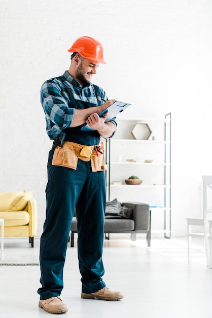 Happy bearded worker in safety helmet writing while holding clipboard in living room