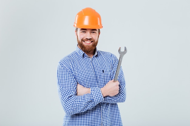 Happy bearded young man in orange helmet standing and holding wrench over white wall