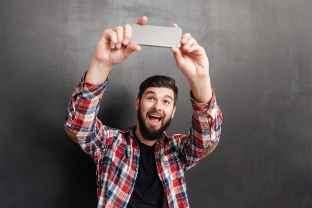 Happy bearded young man in plaid shirt making selfie with mobile phone on grey