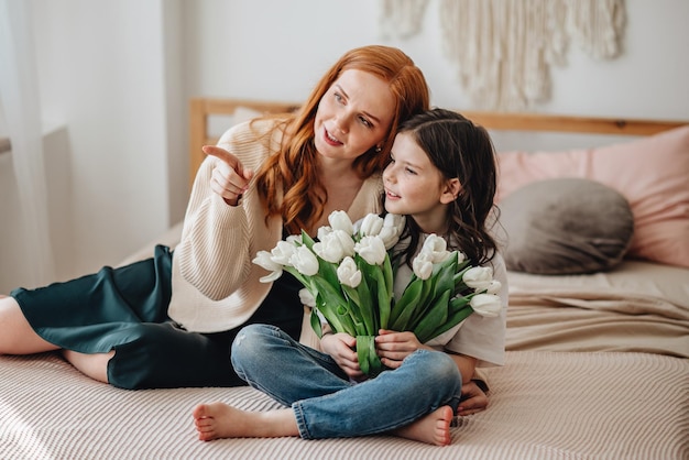 Happy beautiful affectionate mother with a bouquet of spring\
flowers hugs her daughter at home on the bed enjoys the moment of\
surprise celebrates mother\'s day