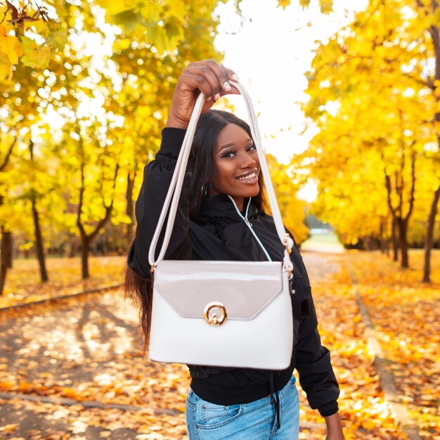 Happy beautiful african black woman with smile in fashionable clothes and jacket showing white leather handbag in park with bright yellow autumn foliage. Female casual style with fashion bag
