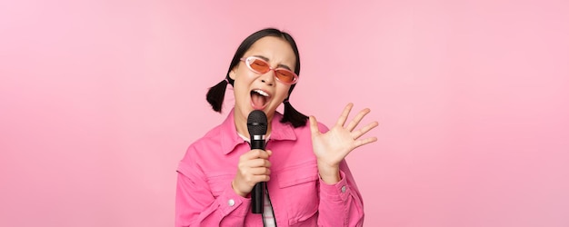 Happy beautiful asian girl singing with mic using microphone enjoying karaoke posing against pink studio background