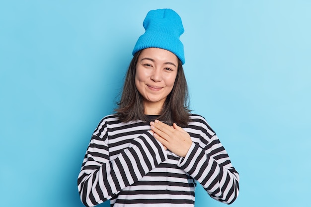 Happy beautiful Asian woman makes gratitude gesture presses palms to heart feels touched pleased by compliments says thank you expresses thankfullness wears blue hat striped jumper poses indoor