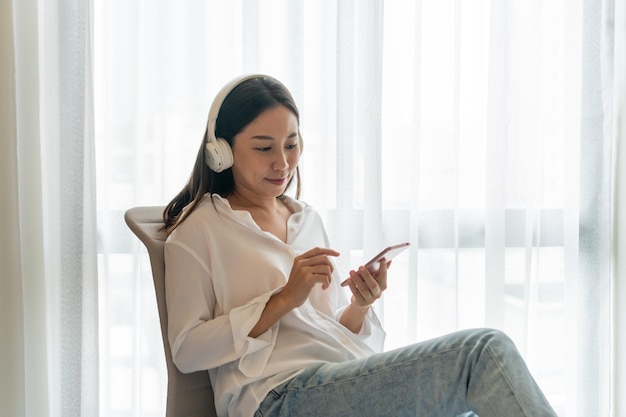 Happy beautiful Asian woman using mobile phone and listening music while sitting at condominium Technology and lifestyle leisure activities on weekend concept Copy space closeup
