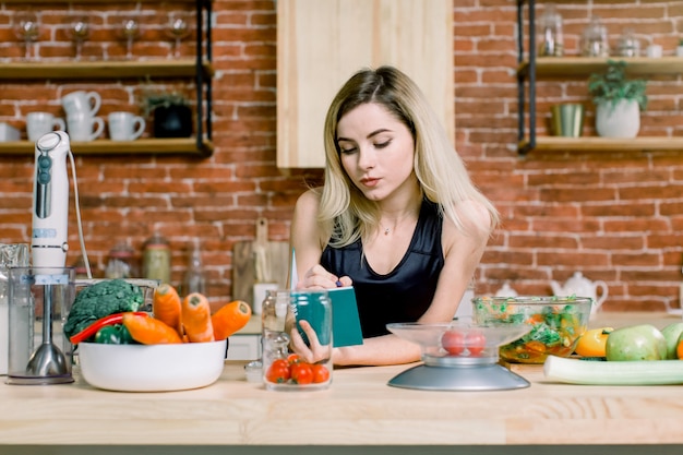 Happy beautiful blond woman standing in her kitchen and writing a shopping list on a notebook at home. Healthy Food - Vegetable Salad. Diet. Dieting Concept. Healthy Lifestyle. Cooking At Home.