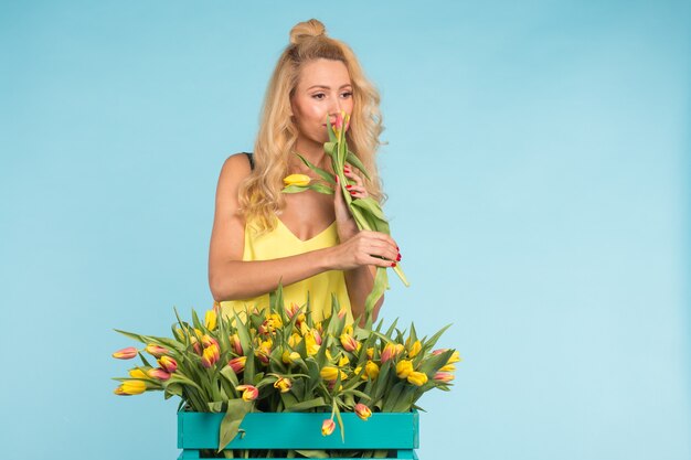 Happy beautiful blonde caucasian woman with big box of tulips on blue surface.