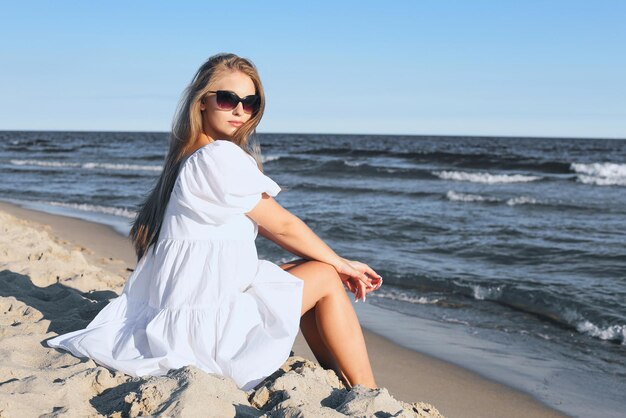 Happy, beautiful blonde woman is sitting on the ocean beach in a white summer dress and sunglasses.