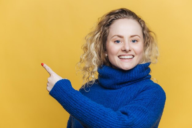 La bella giovane donna bionda felice ha l'espressione soddisfatta indossa un maglione blu casual indica che lo spazio vuoto della copia sul muro dello studio in bianco giallo pubblicizza qualcosa con grande piacere