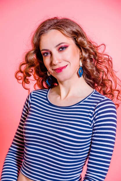 Happy beautiful brunette girl with red makeup, curly hair and a blue stripes sweater posing against a red background. Vertycal photo