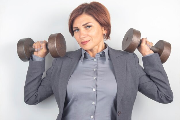 Felice bella donna d'affari in un vestito solleva i manubri su uno sfondo bianco. fitness e salute. crescita professionale e lavoro