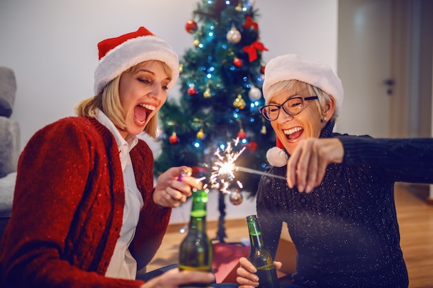 Happy beautiful caucasian blonde woman lightening sparkler and holding beer. Her mother holding sparkler and beer. Both having Santa hats on heard. Family time.