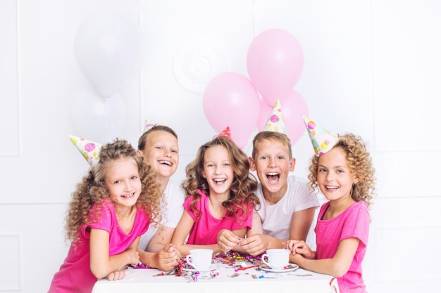 Foto felici bei bambini carini sorridono alla festa di festa con palloncini e coriandoli insieme al tavolo nella stanza bianca