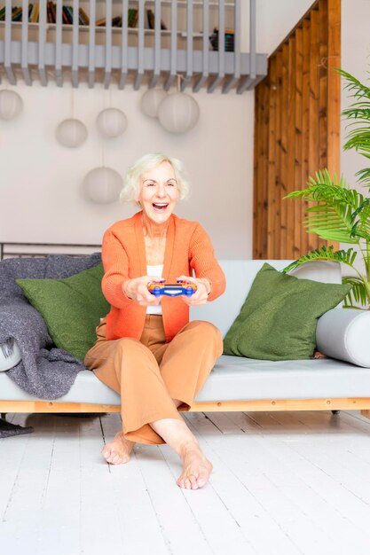 Фото Счастливая красивая пожилая седые волосы пожилая женщина сидит на диване в гостиной уютная квартира улыбается пожилая жена с пультом для игры в консоль современная бабушка играет в видеоигры онлайн