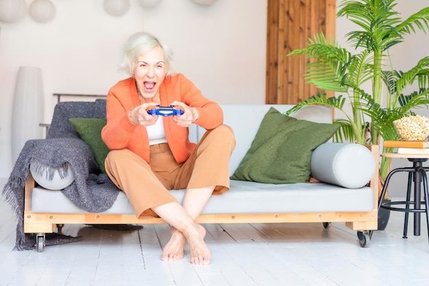 写真 美しい老人灰色のの高齢女性リビングのソファに座って快適なアパートで笑顔の高齢の女性コンソールをプレイするリモコントで現代のおばあちゃんがビデオゲームをオンラインでプレイしています