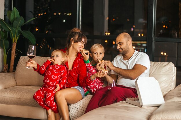 Счастливая, красивая семья вместе ест пиццу на диване в пижаме на рождественские праздники