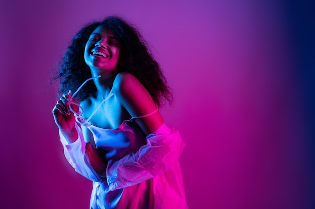 Happy beautiful fashion young african american girl hipster black woman hold stylish glasses wear raincoat look at camera laughing dancing isolated on disco party purple pink trendy studio background