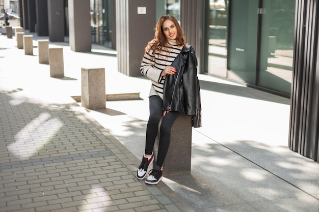 Happy beautiful fashion young girl in fashionable clothes with a leather jacket sweater leggings and stylish sneakers is sitting in the city