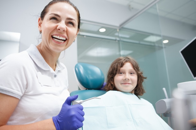 Happy beautiful female dentist laughing while working with child patient at her clinic