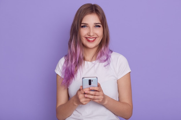 Happy beautiful female wearing white casual t shier holding smart phone in hands, browsing internet or social network, isolated over purple wall.