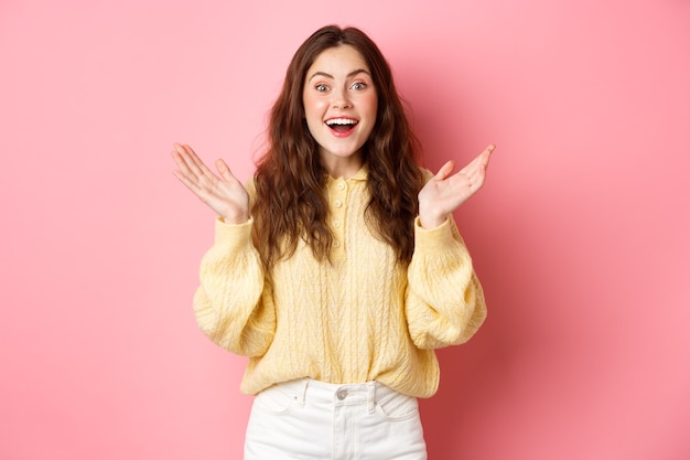 Happy beautiful girl gasping amazed, clap hands and smiling, looking surprised, receive great news, being praised, standing against pink wall.