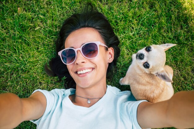 Happy beautiful hipster woman in sunglasses is having fun while she is lying on the lawn and making selfie with her little cute dog