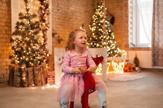Счастливая красивая маленькая девочка в модном платье катается на красных качелях на фоне елки, огней и домашних украшений