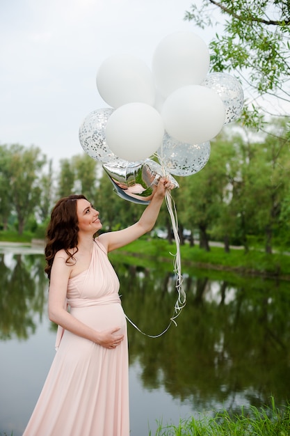 Happy beautiful pregnant woman in pink long dress walking with air balloons in the green p