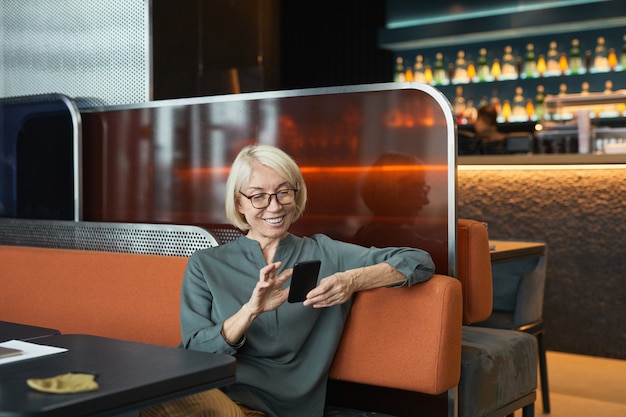 Happy beautiful senior woman with blond hair sitting on sofa and leaning elbow on back of seat while using smartphone for online communication