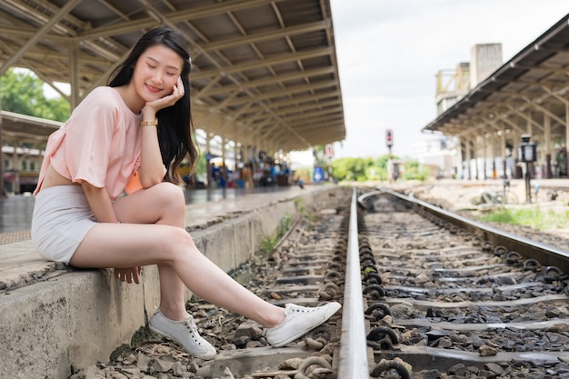 幸せな美しい旅行者は、鉄道の近くに座っている駅で電車を待っている間彼女の目を閉じます。